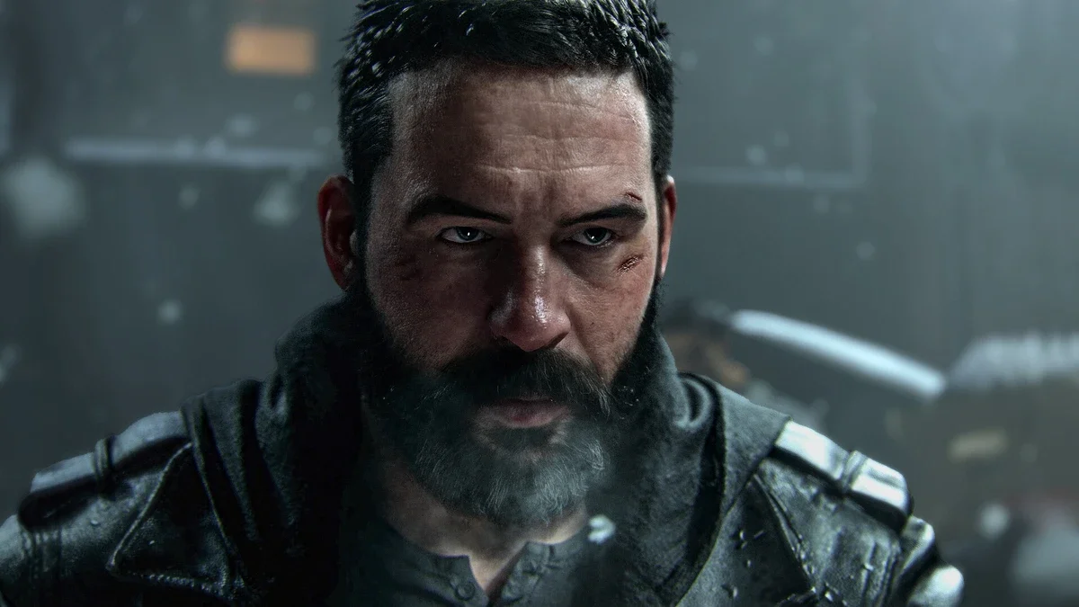 Close-up of a bearded soldier with short dark hair in a dark tactical jacket looking toward the camera in a dim, misty setting