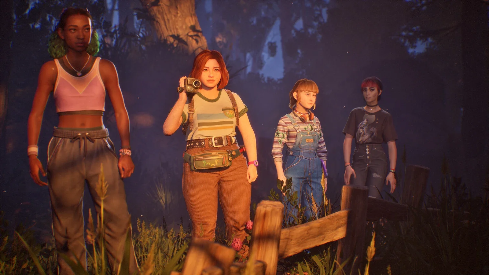 Four teenage girls in 1990s clothes stand by a wooden fence in a dark forest, one holding a camcorder