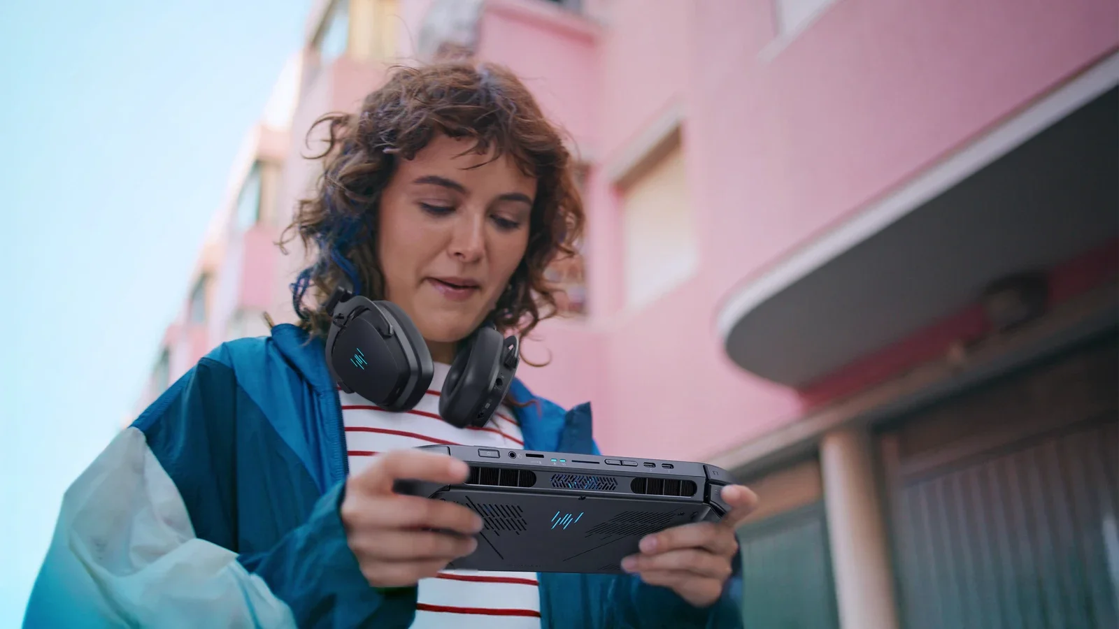 A woman with curly hair and headphones around her neck plays on an Acer Predator Atlas 8 handheld outdoors beside a pink building